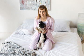 happy woman in pajamas and cute little puppy resting on bed in morning at home