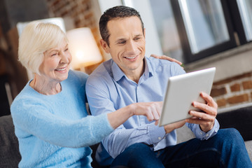 Becoming technology-savvy. Petite senior woman sitting on the couch next to her son and tapping the tablet while her son teaching her how to use it
