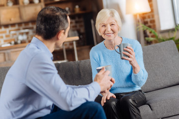 Pleasant conversation. Cheerful senior woman and her young son sitting in the living room, drinking coffee and talking to each other