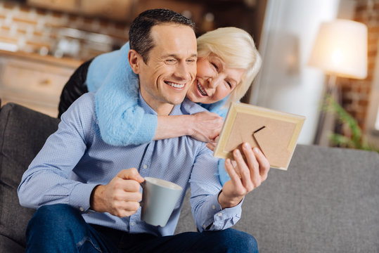 Cozy day. Loving elderly mother hugging her son from behind while he sitting on the couch, drinking coffee and looking at his baby photo in a frame