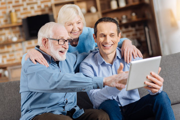 Interesting lessons. Charming senior woman hugging her husband and son sitting on the couch while the younger teaching his father how to use a tablet