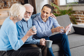 Quality time. Charming young man sitting on the couch next to his senior parents, holding a tablet and chatting with them while they drinking coffee