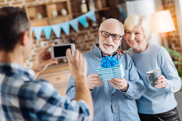 Happy moments of life. Loving young son taking a picture of his elderly parents posing with a birthday present during the party