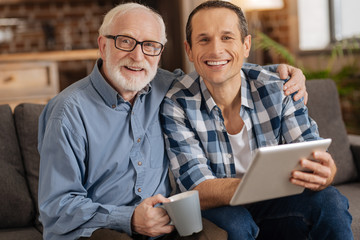 Close relations. Cheerful senior man and his beloved son sitting on the sofa together, bonding to each other and posing for the camera while the young man holding a tablet