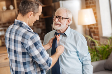 Gentle care. Caring young man tying the tie of his senior father and talking to him while the man...