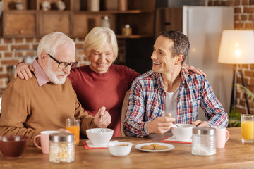 Delicious breakfast. Petite elderly woman hugging her adult son and beloved husband while they sitting at the kitchen counter and eating oatmeal