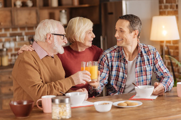 Peaceful morning. Cheerful elderly woman hugging her beloved son and husband while they eating oatmeal and clinking glasses with orange juice