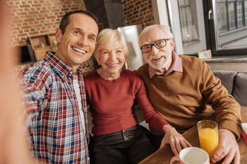 Greatest moments. Upbeat family consisting of an adult son and his senior parents sitting at the kitchen table and taking selfies together