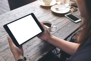 Mockup image of business woman's hands holding black tablet pc with blank white screen and coffee cup on vintage wooden table in cafe background