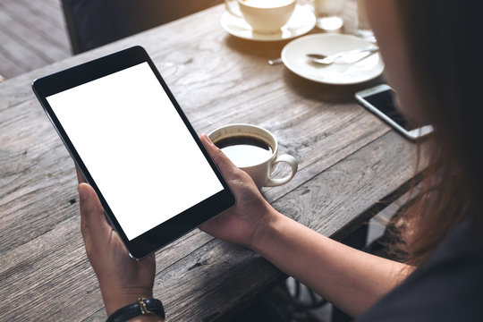 Mockup image of business woman's hands holding black tablet pc with blank white screen and coffee cup on vintage wooden table in cafe background