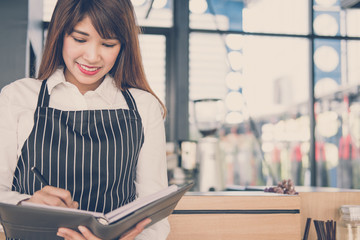 small business owner holding notebook at counter in coffee shop. female barista wearing apron writing note at bar in cafe. food service, restaurant concept.