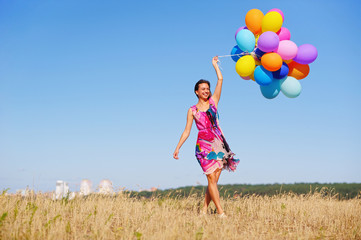 Beautiful, cheerful young woman in a bright dress with colorful balloons  on a field with a blue sky in the summer. Warm sunny summer day with light breeze.