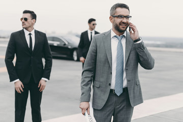smiling businessman walking with bodyguards and talking by smartphone