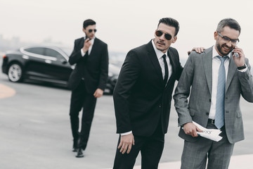 happy businessman walking with bodyguards and talking by smartphone