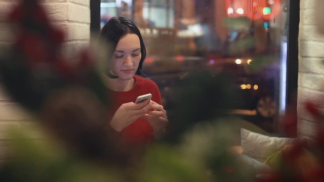 Attractive young woman using smartphone in cafe in evening. Christmas time