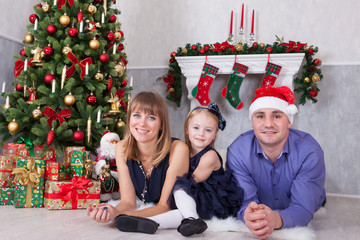 Christmas or New year celebration. Portrait of cheerful happy family of three people lying on the floor near Christmas tree with xmas gifts. A fireplace with christmas stocking behind them