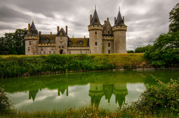 Fototapeta na wymiar Castle of Sully-sur-Loire, Loire region, France. Snap of 30 June 2017 17:38. Shooting from the park towards the castle entrance. The façade is mirrored in the water of the moat. Sky with stormy clouds