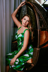 Fototapeta na wymiar Portrait of stylish brown hair woman sitting in green tropical dress, green earrings, with makeup on bamboo armchair in the photostudio