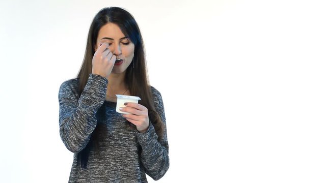 Brunette female eating yoghurt, rasting it and making dislike face, studio shot over white background