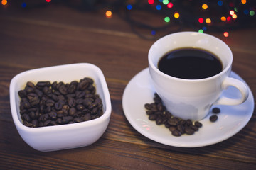 A romantic festive still life with a cup of coffee with a saucer and a bowl of coffee beans on a rustic wooden background. Dark colorful bokeh