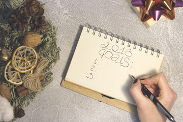 A notebook with 2018 goals inscription, a gift box with an orange-purple shiny bow and hand-decorated pine wreath on a light concrete background. A hand writing in a notebook. Top view