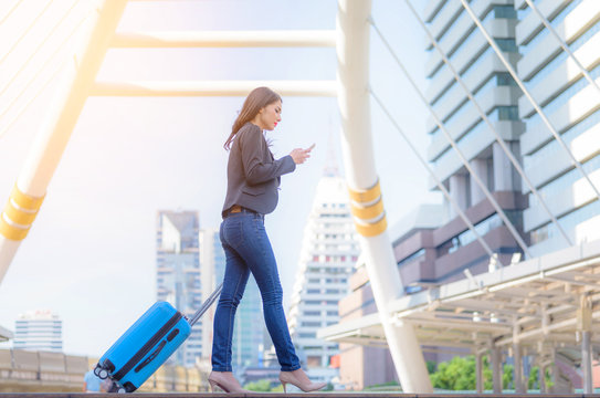 Portrait of business woman looking smartphone with blue travel bag on walkway with blurred city background.