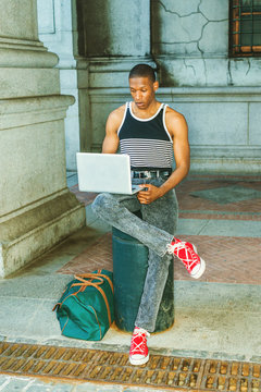 College student traveling, working in New York. Wearing black, white striped tank top, jeans, red sneakers, carrying green bag, a black guy sitting on street, reading, working on laptop computer..