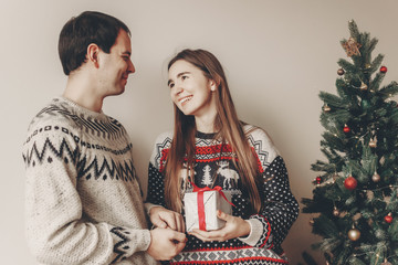 Fototapeta na wymiar merry christmas and happy new year concept. stylish hipster couple in sweaters holding gift with red bow in festive room at christmas tree with lights. happy holidays. family moments