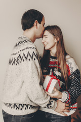 merry christmas and happy new year concept. stylish hipster couple in sweaters holding gift with red bow in room at christmas tree with lights and kissing. happy holidays. family  moments