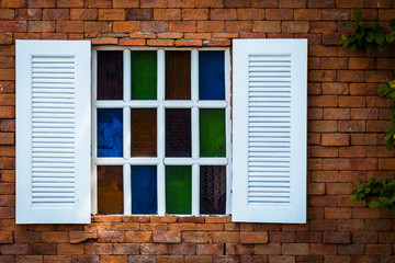 old white wooden window with stained glass on brown brick wall background, stone wall with little colorful stained glass window and green leaf on the right side.