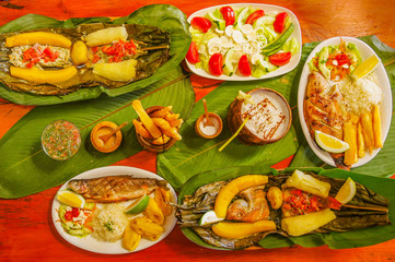 Above view of assorted food, grilled meat with potato, sweet tomatos, salad, pepper, fried plantain, fish in a leaf with yucca, fried fish, fried yucca, palmito salad, served in white plates over a