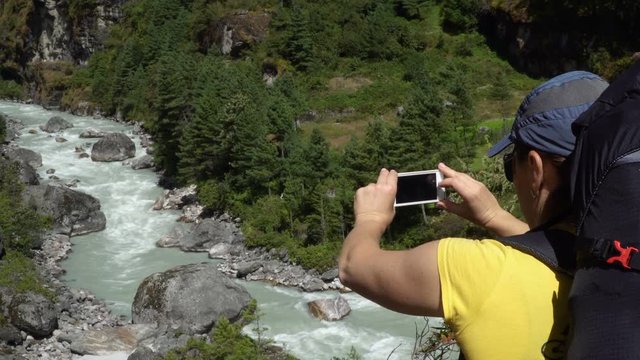 A woman is taking pictures against the background of big mountains and the green mountain river. Enjoys adventure and travel concept