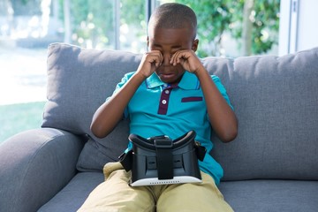 Boy rubbing eyes while sitting with VR headset on sofa