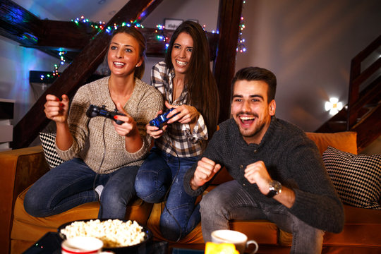 Two female best friends sitting at home on pleasant  evening and playing games on console.They challenge each other to win while man cheering.