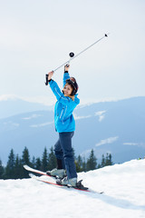 Cheerful woman skier wearing blue ski suit and black helmet, holding poles above a head, smiling to...