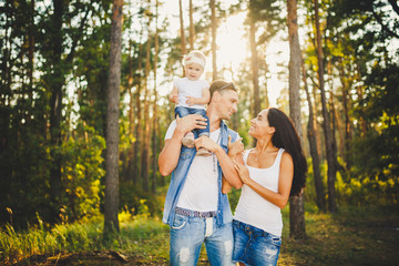 stylish young Family of mom, dad and daughter one year old blonde sitting near father on shoulders, outdoors outside the city in a park amid tall trees in summer at sunset.
