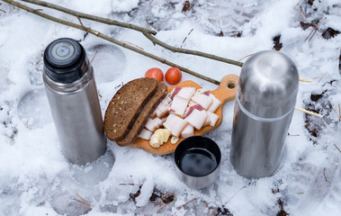 lard, bread, with tomatoes on a wooden cutting board in the snow next to the thermos with tea.