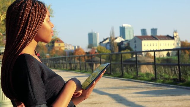 Young black woman sits on the bench and works on tablet - city in the background