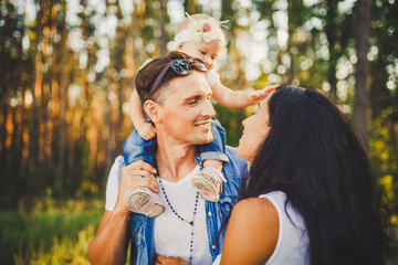 Fototapeta premium stylish young Family mom, dad and daughter one year old blonde sitting with father on shoulders playing happy and smiling, outdoors outside the city in the woods in summer at sunset.