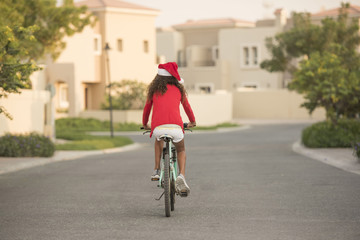 Naklejka premium A biracial child wearing Santa hats while riding her bike on a street in a neighbourhood with homes in the background