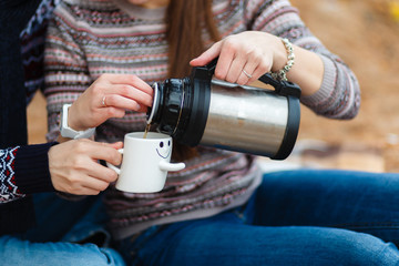 Young couple drinking hot tea from a thermos and pouring it into white circles, trying to stay warm. A guy and a girl in jeans and sweater, sitting on a blanket in the fresh air, enjoying nature.