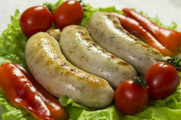 Ready-fried sausages for the grill are on the green leaves of lettuce next to tomatoes and slices of sweet pepper. Close-up.