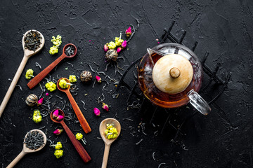 Brew the aromatic tea. Tea pot near wooden spoons with dried tea leaves, flowers and spices on black wooden background top view