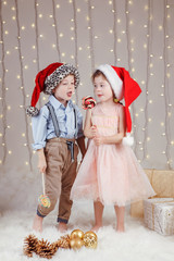 Portrait of white Caucasian children friends wearing Santa Claus hats, celebrating Christmas or New Year. Little cute girl and boy eating lollipops in studio with winter holiday decoration