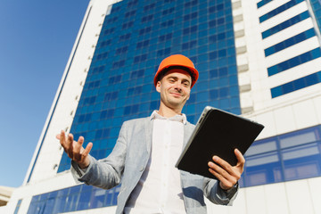 Successful business man in gray suit and protective construction orange helmet with tablet on background of building in blue white color. Engineer in helmet for workers security on office buildings.