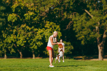Mother and daughter throwing orange flying disk to small funny dog, which catching it on green...