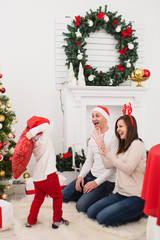 Happy young cheerful parents fighting pillows with cute little son. Child boy in red hat in light room at home with decorated New Year tree. Christmas good mood. Family, love and holiday 2018 concept.