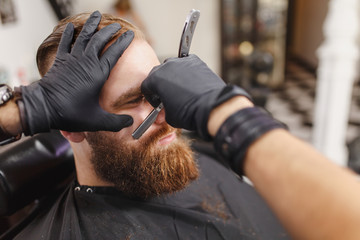 Close up male professional hairdresser serving client, shaving thick big beard straight razor....