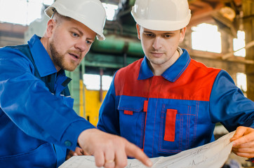 Workers in overalls and white protective helmets view the drawing on the background of an industrial metal plant. Builders in shape. Men are engineers