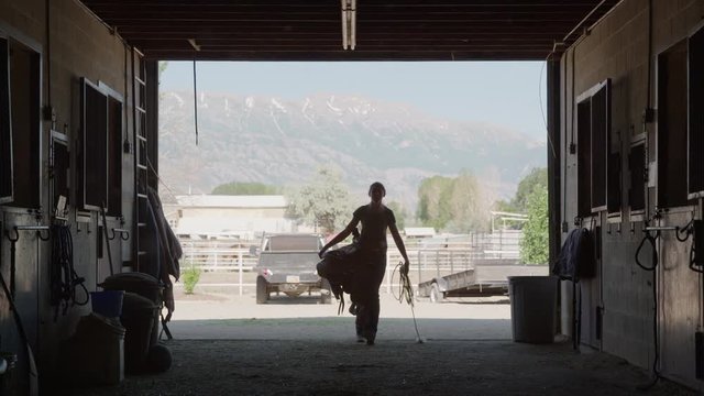 Wide slow motion silhouette of girl carrying saddle and rope into stable / Lehi, Utah, United States